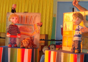 Po lewej stronie animatorka trzyma marionetkę chłopca i psa, po prawej marionetkę drugiego chłopca.